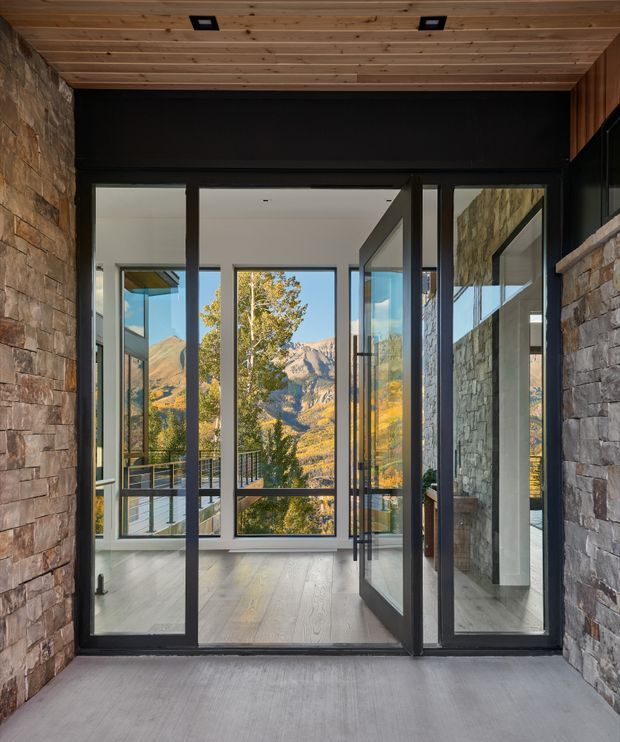 A modern glazed entry or transition space framed by rough stacked-stone walls, with a warm timber-lined ceiling and dark metal-framed full-height glass doors and windows. The open doorway leads into a bright interior with pale timber flooring, while mountain scenery and autumn trees are visible beyond the glazing. The composition emphasises strong indoor-outdoor connection, natural stone, timber, and expansive views.