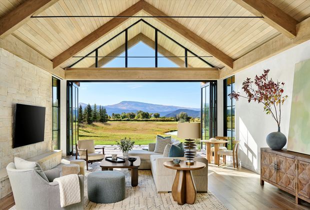 Bright, spacious living room with a dramatic timber-lined pitched ceiling, exposed structural beams, and a large glazed gable opening onto expansive rural and mountain views. The room features light stone walls, pale timber flooring, contemporary upholstered seating, a television, sculptural coffee tables, a dining area, and a patterned timber sideboard. Open glass doors create a strong indoor-outdoor connection.