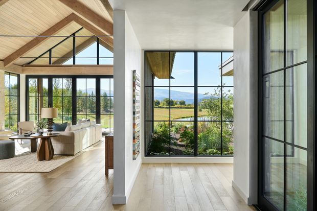 A bright contemporary interior with a glazed living room opening onto expansive rural views. The living area has a pitched timber-lined ceiling with exposed structural beams, extensive black-framed glazing, pale timber flooring, and a neutral seating arrangement. A connected glazed transition space or entry zone occupies the foreground, with landscaped planting visible outside.