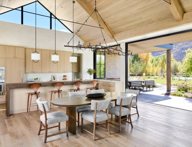 Bright open-plan kitchen and dining area with a large timber-lined vaulted ceiling, exposed timber beams, pale wood cabinetry, a stone island with bar seating, and a round dining table with upholstered chairs. Black-framed glazing and wide open doors connect the interior to a covered outdoor dining terrace and landscaped garden. A sculptural dark metal chandelier hangs over the dining table, while pendant lights illuminate the island.