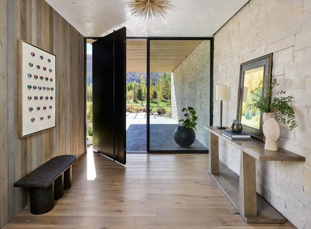 A refined modern entry hall with a sculptural black pivot front door opening to a landscaped exterior courtyard. The space features full-height glazing, warm vertical timber wall panelling, wide-plank timber flooring, a pale textured stone feature wall, and a minimalist console with contemporary decorative objects. A dark upholstered bench and framed artwork sit along the timber wall, while a starburst pendant adds a subtle focal point overhead.