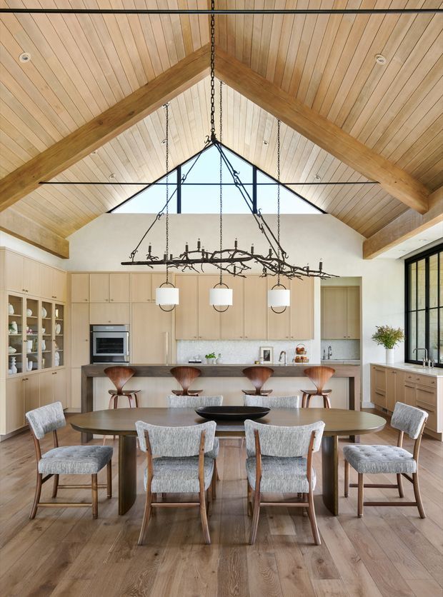 A bright, open-plan kitchen and dining space beneath a dramatic timber-lined pitched ceiling with exposed beams and a triangular clerestory window. The kitchen features pale wood cabinetry, a light stone benchtop and splashback, integrated appliances, and a long island with timber bar stools. In the foreground is a large oval timber dining table surrounded by upholstered dining chairs, with a sculptural branch-like chandelier suspended above.