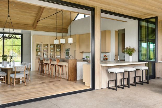 A bright open-plan kitchen and dining area opening onto a covered outdoor entertaining space through large black-framed sliding and folding glass doors. The interior features extensive light timber lining on the ceiling and floors, light timber cabinetry, a kitchen island with seating, pendant lights, and a dining table beside full-height glazing. The covered exterior includes a matching timber-clad outdoor kitchen or bar with stools.