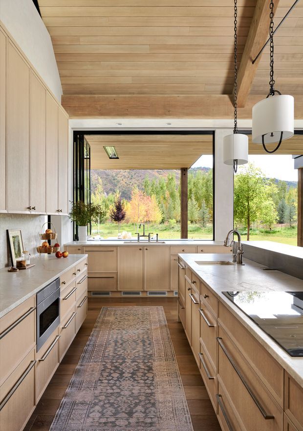 A warm, light-filled contemporary kitchen with extensive pale timber cabinetry, a large stone island with integrated sink, and matching perimeter counters. The galley-style layout opens to expansive black-framed glazing overlooking a green, wooded landscape. A timber-lined ceiling with exposed structural beams, pendant lights suspended from chains, integrated appliances, and a patterned runner complete the inviting space.