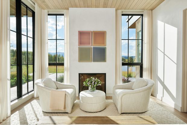 Bright, serene sitting room with two cream boucle lounge chairs and a matching round ottoman arranged around a central fireplace. The room features floor-to-ceiling black-framed glazing, a pale timber-lined ceiling, light neutral walls, a textured area rug, and views across a landscaped rural setting.
