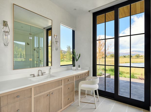Bright, refined bathroom with a long double-sink vanity in light timber joinery and a white countertop. A large framed mirror reflects a glass-enclosed shower, while black-framed floor-to-ceiling glazed doors open to a scenic landscape and water view. The space has pale surfaces, contemporary fixtures, decorative wall sconces, and a sculptural white stool.
