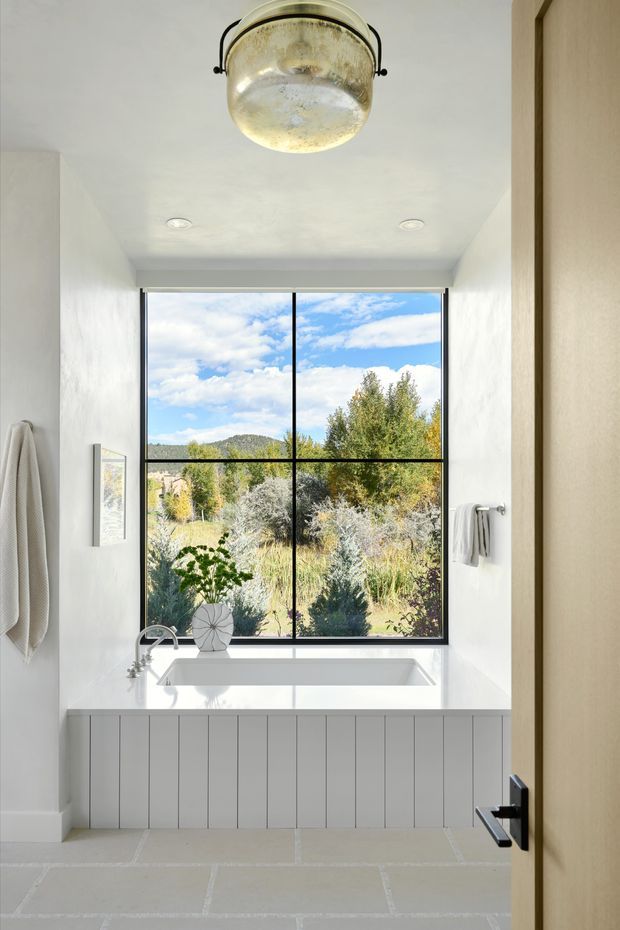 Bright, serene bathroom viewed through a partially open timber door, featuring a freestanding white bathtub beneath a large black-framed four-pane window. White textured walls, white vertical-panel bath cladding, pale stone-look floor tiles, a wall-mounted towel rail, and a vintage brass-toned pendant light create a refined modern country feel. The window overlooks landscaped vegetation and distant hills.