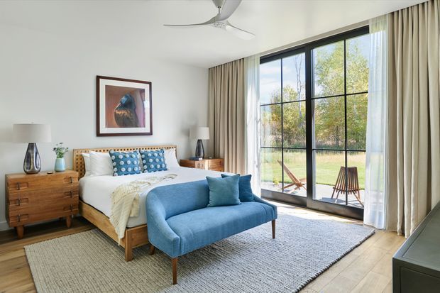Bright, well-appointed bedroom with a timber bed frame and matching mid-century-style chest of drawers and bedside tables. The bed is dressed in white linen with patterned blue cushions, complemented by a blue upholstered bench at the foot. Large black-framed floor-to-ceiling glazed doors open toward a garden, with full-length beige curtains. A neutral textured rug, timber flooring, ceiling fan, bedside lamps, and framed artwork complete the calm, contemporary space.