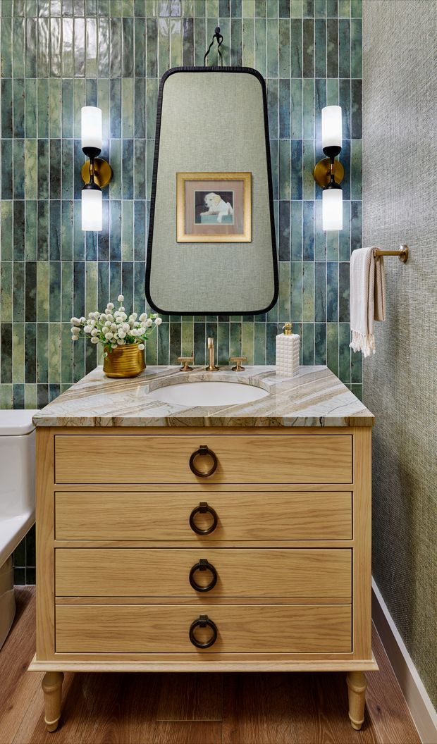 A richly detailed bathroom vanity vignette featuring a freestanding light-wood drawer vanity with dark circular pulls, a veined marble benchtop and integrated basin, and a large rounded rectangular mirror. The wall behind is clad in vertically arranged glossy green-blue rectangular tiles, flanked by sculptural wall sconces. A brass towel rail, framed artwork, flowers, and bathroom accessories add decorative character. The room has a warm, refined, vintage-inspired feel.