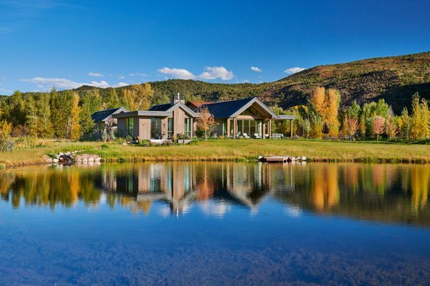 A low-profile contemporary house set beside a tranquil pond, surrounded by autumnal trees and open lawn, with a wooded hillside rising behind it. The home has dark pitched roof planes, extensive glazing, timber-toned wall sections, and a strong reflection in the water. The scene is a bright, serene landscape-oriented exterior view.