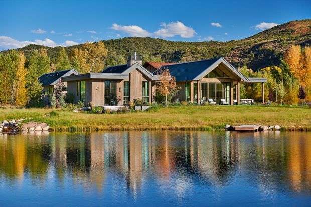 A contemporary single-storey residence set beside a reflective pond in a wooded mountain landscape. The home features multiple pitched gables, dark metal roofing, timber-clad wall sections, extensive glazed openings, and a broad sheltered outdoor terrace. Autumn planting and forested hills surround the property.