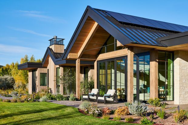 Exterior view of a contemporary single-storey residence with multiple pitched gables, broad roof overhangs, dark standing-seam metal roofing, timber-clad soffits and exposed timber posts. Large floor-to-ceiling glazed openings connect the living spaces to a landscaped garden and covered outdoor sitting area. A chimney, solar panels, stone-clad wall sections and carefully planted garden beds are visible under clear daylight.