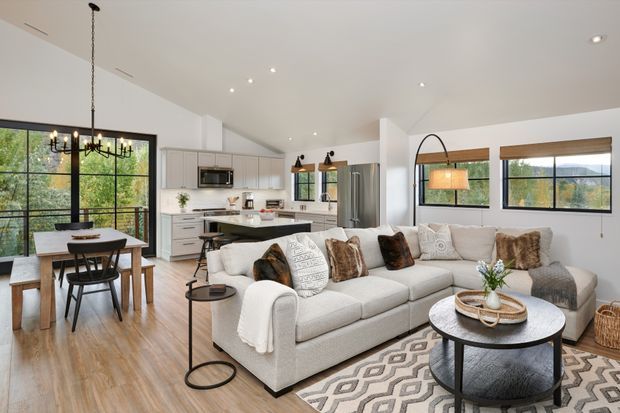 Open-plan contemporary living and dining area with a visible kitchen in the background. The room features a large light fabric sectional sofa, round coffee table, patterned rug, timber flooring, black-framed windows, a dining table with pendant chandelier, and a neutral white and grey palette with warm wood accents.
