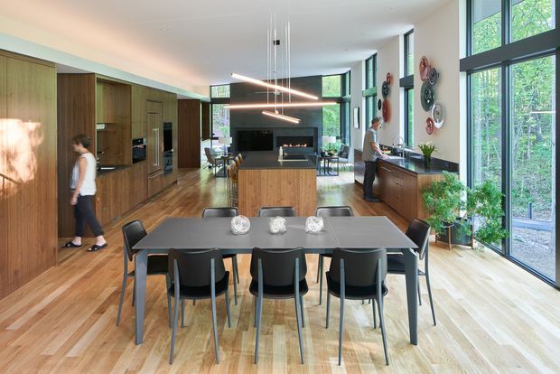 Open-plan contemporary kitchen and dining area with extensive warm timber cabinetry and flooring, a central kitchen island, a dark dining table with black chairs, and a linear fireplace feature in the background. Floor-to-ceiling black-framed glazing brings in abundant natural light and views of trees. Decorative wall objects, pendant lighting, and indoor plants complete the refined modern interior.