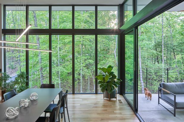 A contemporary dining and living area enclosed by expansive floor-to-ceiling glazing, with dark-framed window walls overlooking a dense wooded landscape. A long dining table with pendant lighting occupies the foreground, while a glazed opening leads to an outdoor balcony or terrace furnished with a lounge chair. Light timber flooring, dark structural framing, and indoor greenery create a bright, nature-connected interior.