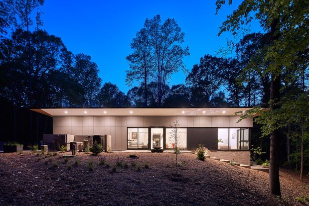 A low, single-storey contemporary pavilion-style home set within a wooded landscape, photographed at dusk. The building has a long horizontal form, a flat roof with recessed downlights, extensive floor-to-ceiling glazing, and pale panelled exterior walls. A dark gravel or mulch forecourt with sparse landscaping leads to the illuminated interior.