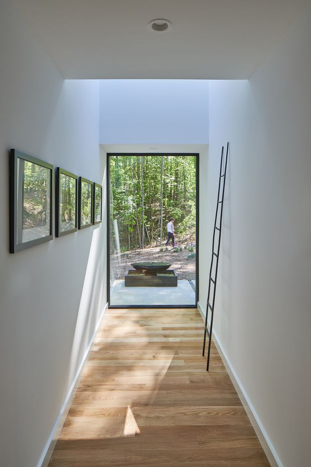 A narrow, minimalist interior passage with white walls and ceiling, a light timber floor, framed artwork along the left wall, and a tall black-framed glazed door or window at the end opening toward a wooded landscape. A slender black ladder-like sculptural element leans against the right wall. Natural daylight creates strong, elongated shadows across the floor.