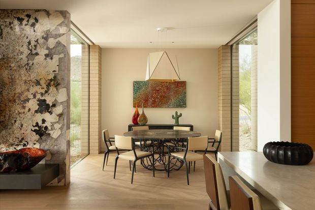 A refined dining area with a large rectangular table and upholstered chairs, framed by full-height glazing and warm timber-lined walls. A dramatic natural-stone fireplace feature occupies the left side, while sculptural artwork and decorative objects add visual interest. The room has a warm, contemporary character with abundant natural light and an open connection to adjacent spaces.
