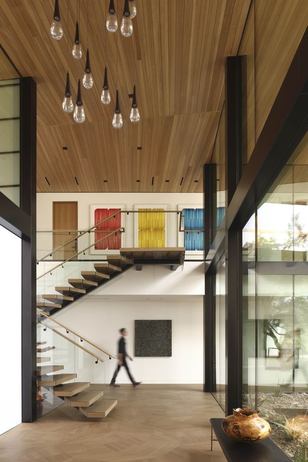 A refined double-height interior hall or entry space with a floating timber staircase, glass balustrades, dark structural steel framing, expansive glazing, and a warm timber-lined ceiling. Multiple suspended pendant lights hang above the stair and central circulation area. The lower level has a herringbone timber floor, while colourful framed wall artwork and a console add visual interest. The space is bright, open, and contemporary, with a mezzanine or upper landing visible.