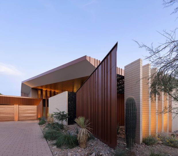 Contemporary residential facade viewed from the driveway, featuring a dramatic angular roof or canopy, tall rust-toned vertical metal screening, pale horizontal masonry walls, warm recessed lighting, and desert-style landscaping with gravel and sculptural cacti. A timber garage or entry gate and paved driveway are visible in the foreground.