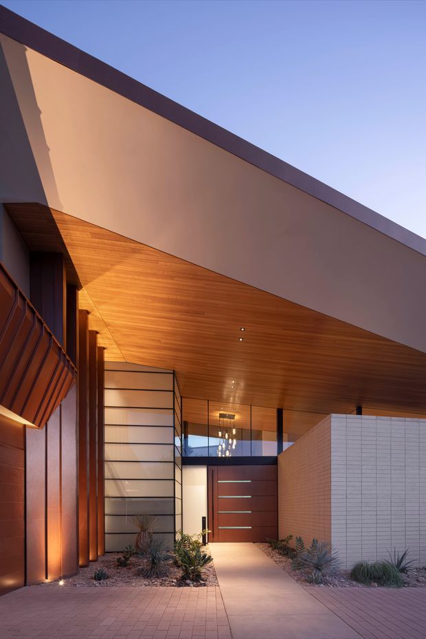A dramatic contemporary residential entrance beneath a large cantilevered roof canopy. The facade combines warm timber soffit cladding, dark metal framing, glazed panels, a tall timber entry door, and a pale tiled feature wall. A paved pathway leads through drought-tolerant landscaped beds toward the entry, with strong angled shadows and warm architectural lighting creating a refined evening atmosphere.
