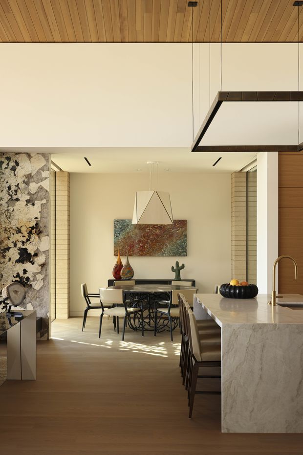A refined open-plan kitchen and dining area with warm timber ceiling and flooring, a large pale stone island with waterfall edge, and dark wood cabinetry. The dining area features a sculptural pendant light, artwork, and a dark dining table with modern chairs. A dramatic stone-clad wall and framed opening add texture, while a large geometric suspended light and recessed ceiling lighting contribute to the contemporary, high-end interior.