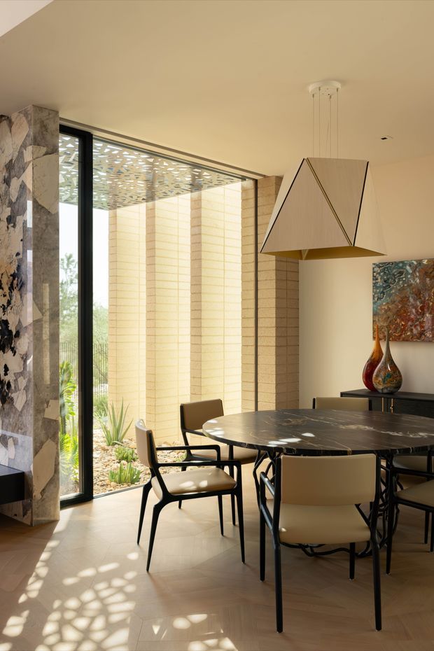 A refined dining room with a large dark stone oval dining table, sculptural upholstered dining chairs, and a geometric pendant light. Full-height black-framed glazing opens onto a landscaped courtyard with pale masonry screening walls, while a dramatic stone-clad column and abstract artwork add visual interest. Warm natural light creates patterned shadows across the timber floor.