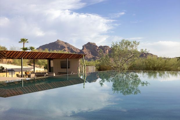A tranquil exterior view of a modern pool area with a broad reflecting pool in the foreground. A low open-sided pavilion with a slim dark metal roof and outdoor seating sits beside the pool, while desert vegetation and rugged rocky mountains form the background. The scene has strong reflections, warm natural light, and a resort-like atmosphere.