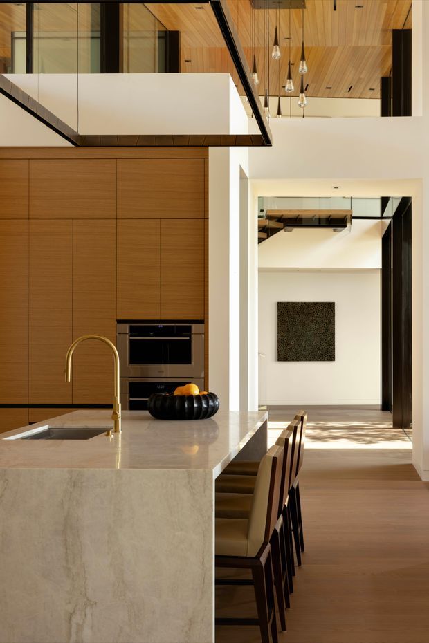 A refined open-plan kitchen interior with a large pale stone island, integrated sink and brushed gold-toned tap, extensive warm timber cabinetry, and built-in black appliances. The kitchen opens to a dining and living area with timber flooring, a dramatic double-height void, glass balustrades, and a timber-lined ceiling featuring multiple pendant lights. Strong natural light enters the adjoining space, creating a warm, contemporary atmosphere.