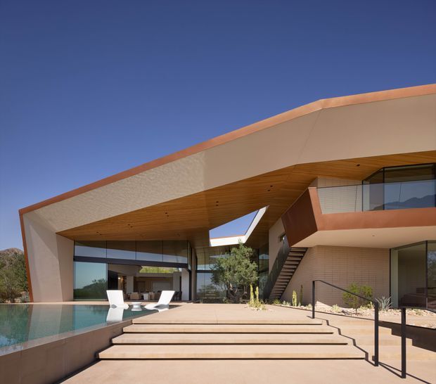 Exterior view of a large contemporary residence with a dramatic cantilevered and sloping roof, extensive floor-to-ceiling glazing, warm timber soffits, terracotta-toned trim, and an elevated terrace accessed by broad steps. A swimming pool and outdoor seating are visible in the foreground, with an open-plan interior glimpsed through the glazing.