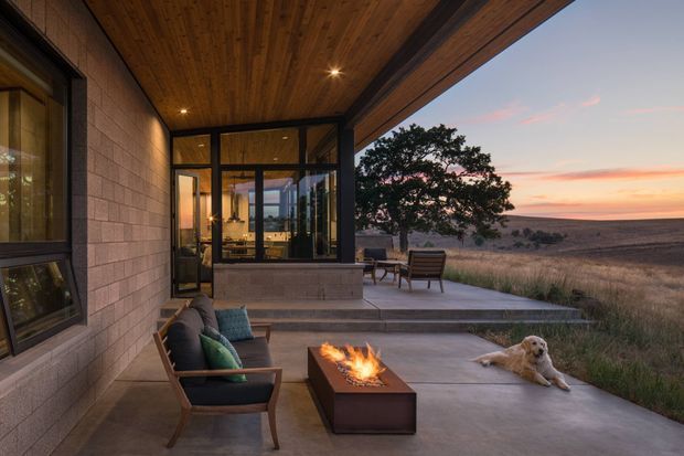 A modern, single-storey indoor-outdoor living area opening onto a broad concrete terrace overlooking an open rural landscape at sunset. The covered terrace has a warm timber-lined ceiling with recessed lighting, dark-framed glazing and doors, exposed concrete block walls, outdoor lounge chairs, a low rectangular fire table, and a dog resting beside it. The glazed interior behind the terrace appears to contain a kitchen or living space.