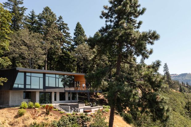 A modern multi-level house set into a wooded, sloping site. The facade combines extensive floor-to-ceiling glazing, dark vertical cladding, exposed concrete walls, and warm timber decking, with broad roof overhangs and a terrace overlooking the surrounding forested landscape.