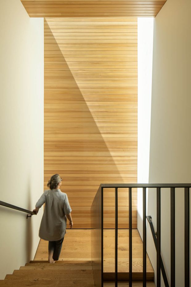 A modern interior stairway descending toward a landing, with warm timber flooring and extensive horizontal timber wall and ceiling lining. A tall sunlit timber feature wall is framed by pale plaster walls, while black metal balustrades and handrails provide strong contrast. A person is walking down the stairs. The composition is minimalist, geometric, and warm, with dramatic natural light and shadow.