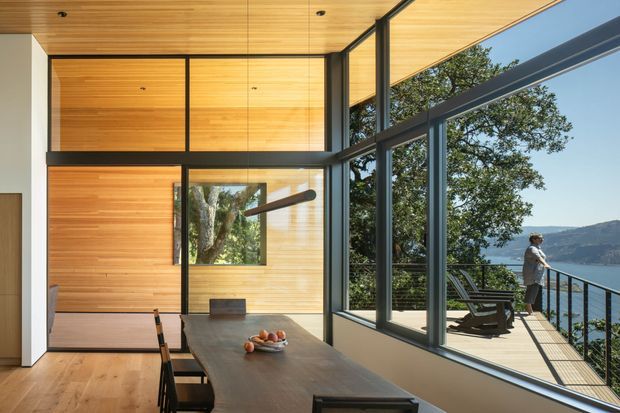 A bright, double-height contemporary dining and living space with extensive floor-to-ceiling glazing overlooking a wooded, mountainous waterfront setting. Warm timber wall and ceiling lining contrasts with slim dark window frames, while a long timber dining table extends toward a deck with outdoor seating.