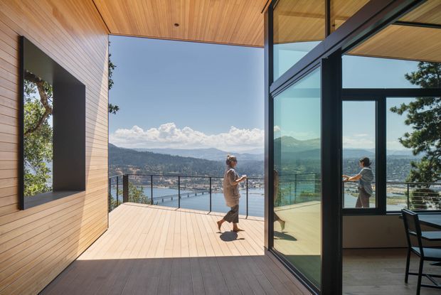 Contemporary timber-lined indoor-outdoor living space opening onto a spacious elevated deck, with expansive black-framed glazing and views across a lake and mountainous landscape. A person stands on the deck, while the glazing reflects the interior dining area. The design emphasizes warm natural timber, strong indoor-outdoor connection, and panoramic views.