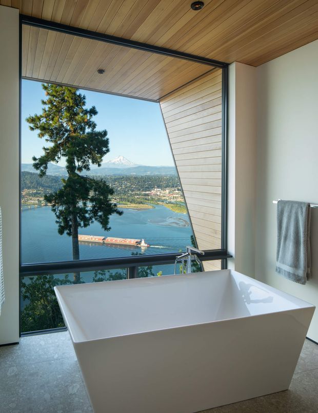 A contemporary bathroom with a freestanding white bathtub positioned beside expansive floor-to-ceiling glazing. The room has a warm timber-lined ceiling and angled timber reveal around the window, dark metal window framing, pale walls, and a grey tiled floor. A towel hangs on a wall-mounted rail. The glazing offers a dramatic view over a lake, town and distant mountain.
