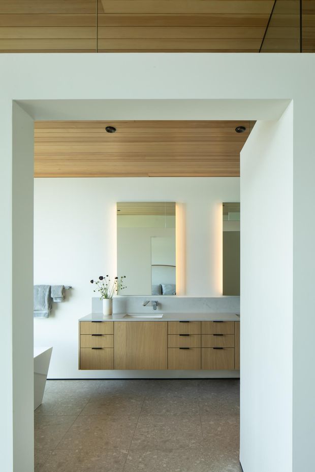 A minimalist contemporary bathroom viewed through a wide doorway, featuring a floating light-wood vanity with dark pulls, a pale stone benchtop, integrated basin and wall-mounted tap, large illuminated mirrors, white walls, and a warm timber-lined ceiling recess. The floor is finished in large-format grey-brown stone or concrete-look tiles.