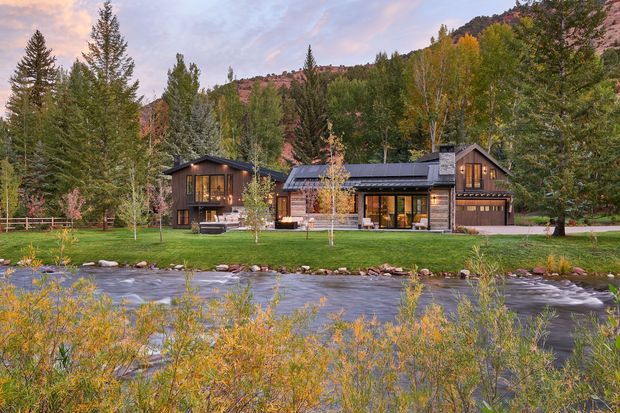 A single-storey timber-clad mountain residence viewed across a flowing river. The home has multiple pitched gabled roof forms, dark-framed glazing, warm interior lighting, a covered entry or outdoor living area, and an integrated garage. It sits in a landscaped grassy clearing surrounded by dense coniferous and autumnal woodland, with a rocky mountain slope behind.