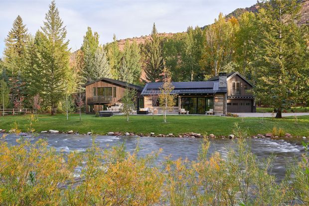 A contemporary timber-clad mountain residence viewed across a flowing river, set within a landscaped forested site. The low-slung main pavilion has a dark pitched roof, extensive glazing, a covered entry or living area, and a connected taller gabled wing with an integrated garage. Autumn trees and wooded hills surround the property.