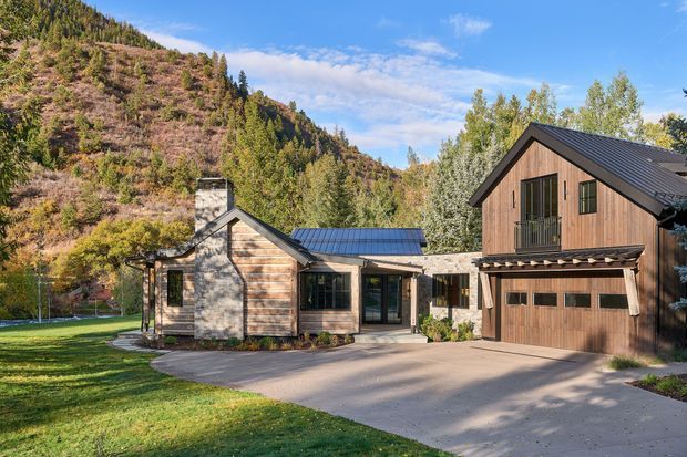 Exterior view of a contemporary mountain home composed of interconnected gabled volumes. The house features natural timber cladding, dark standing-seam metal roofs, stonework around the central chimney and entry, large glazed doors and windows, and an integrated double garage. A paved driveway and lawn sit in front, with wooded hills rising directly behind the property.