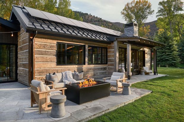 Exterior view of a compact lodge-style home set in a wooded, mountainous landscape. The building has rustic horizontal timber-log walls, a dark metal gable roof with roof-mounted solar panels, dark-framed glazing, and a stone chimney. A covered patio extends along the front, furnished with outdoor lounge chairs around a rectangular fire feature, with stone paving and lawn surrounding it.