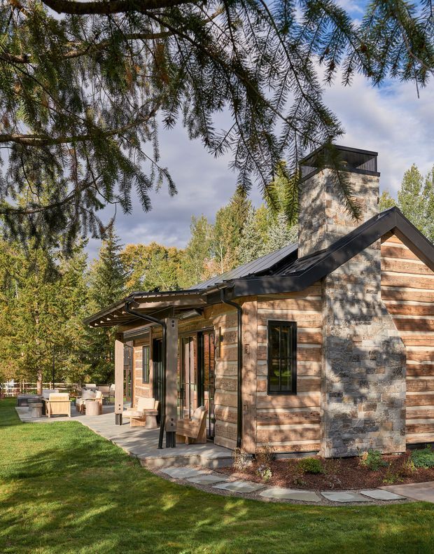 Exterior view of a compact rustic contemporary cabin or residence set within a landscaped, wooded site. The building features horizontal weathered timber cladding, a prominent irregular natural-stone chimney tower, dark metal roofing, black-framed windows and doors, and a covered patio or entry terrace. Mature trees, lawn, stepping-stone paving and outdoor seating surround the structure.