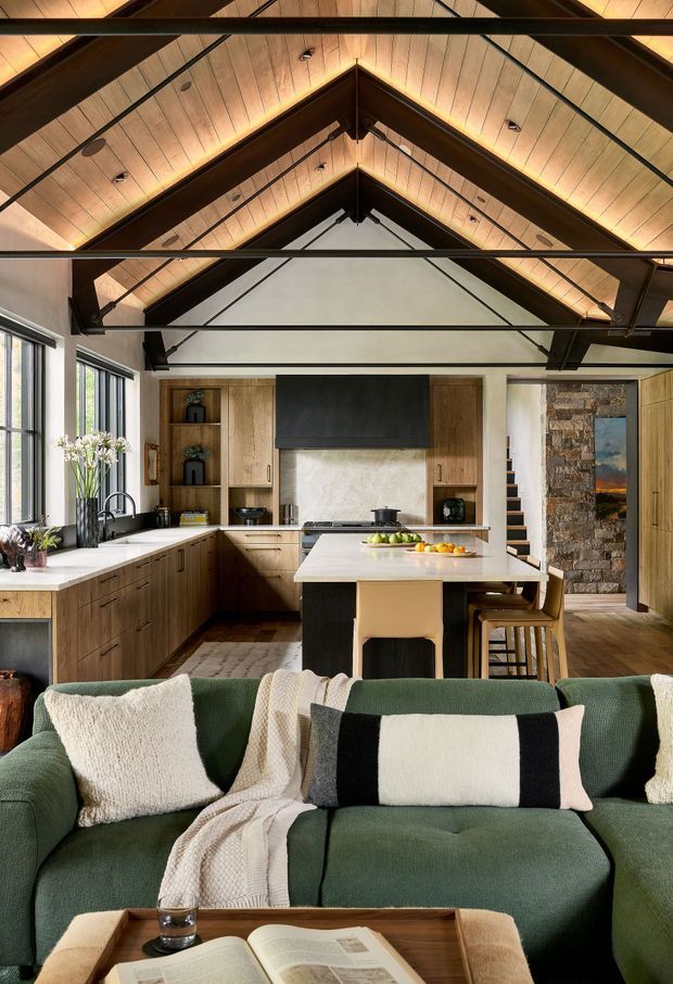 A warm open-plan kitchen and living room with a prominent timber-lined vaulted ceiling and exposed dark structural beams. The kitchen features extensive mid-tone timber cabinetry, pale stone benchtops, a central island with seating, and a stone-clad wall beside a stairway. In the foreground is a deep green sectional sofa with cream and black-and-white cushions and a timber coffee table, creating a comfortable contemporary-rustic interior.