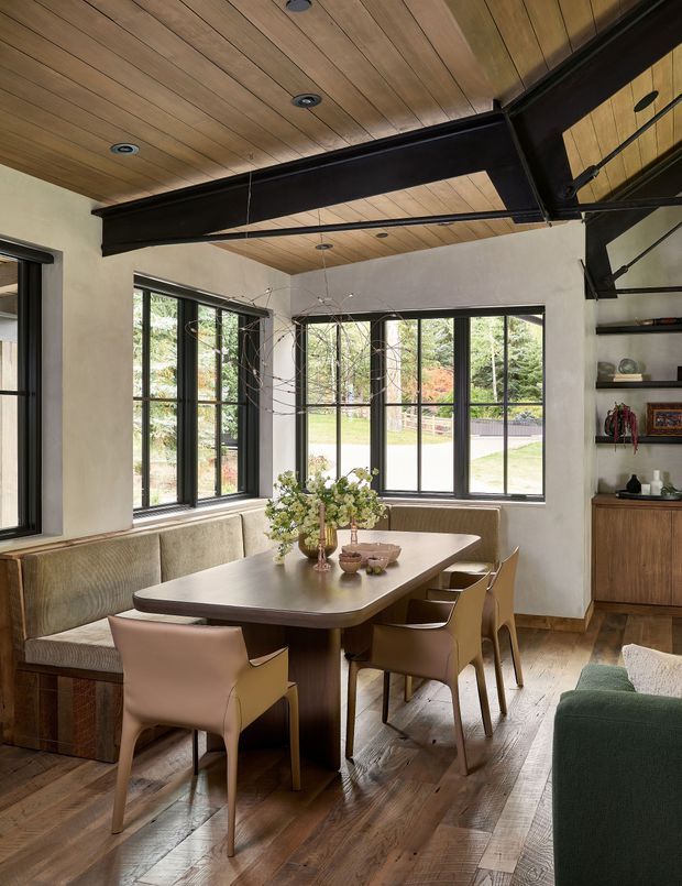 A warm, refined dining area with a large timber dining table, upholstered built-in banquette, and sculptural dining chairs. The room features dark exposed structural beams, a timber-planked ceiling with recessed lighting, plastered walls, dark-framed expansive windows, and timber flooring. Built-in shelving and cabinetry are visible along the right wall, while part of a living room sofa appears in the foreground.
