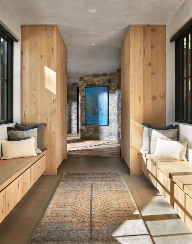 A narrow, symmetrical entry hall or passage with built-in timber bench seating and tall timber storage cabinetry on both sides. Large black-framed windows bring in strong sunlight, while a rough stone feature wall and blue artwork are visible at the far end. The space has a warm, natural, contemporary feel with a neutral tiled floor and patterned runner.
