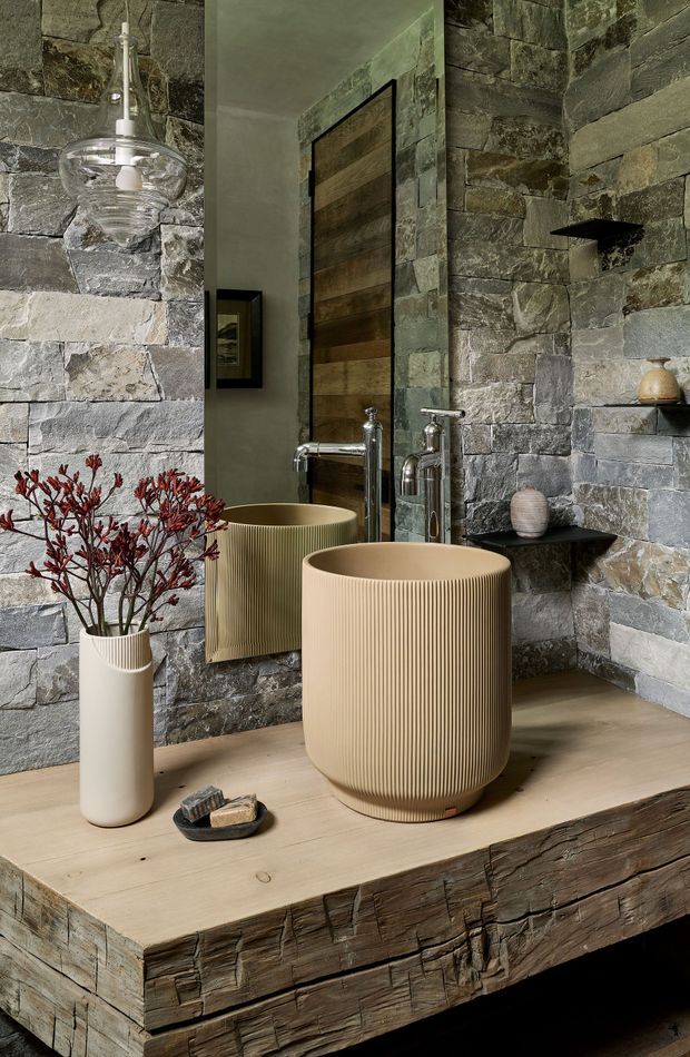 A moody, richly textured bathroom vanity featuring a vessel basin with vertical fluting, polished chrome wall-mounted tapware, a large mirror, and a thick rustic timber countertop with a live-edge-like textured front. The walls are clad in irregular grey natural stone, complemented by a dark timber door and small black wall shelf. Neutral ceramic accessories and a tall vase with red branches add restrained styling.