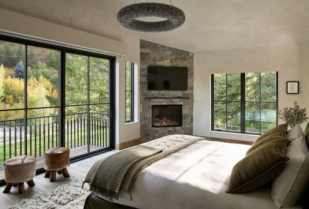 A bright, spacious bedroom with large black-framed windows and glazed doors opening toward a leafy outdoor setting. The room features a textured stone fireplace wall with a television above the firebox, a large upholstered bed with layered neutral bedding, a textured area rug, occasional stools, and a sculptural circular pendant light. Natural materials and abundant greenery views create a warm, refined retreat.