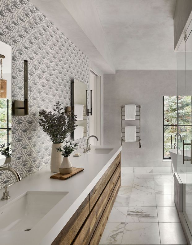 A bright, elongated bathroom featuring a long double-sink vanity with a white solid-surface countertop and warm timber drawer fronts. The left wall has patterned botanical wallpaper, while the far wall is finished in smooth pale grey. Large-format white marble-look floor tiles, frameless shower glazing, chrome fixtures, heated towel rails, mirrors, and black-framed windows create a refined contemporary interior.