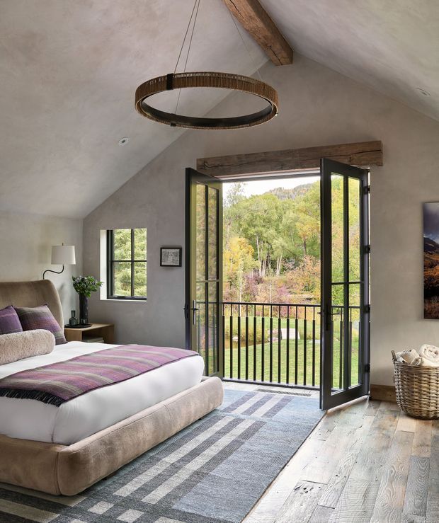 A warm, rustic bedroom with a sloped vaulted ceiling and exposed timber beam. The room contains a large upholstered bed, bedside table, floor lamp, patterned throw, striped rug, and woven basket. Black-framed glazed French doors open to a small balcony with a black metal balustrade and views of leafy woodland. The interior uses plastered walls, pale timber flooring, and a circular woven pendant light.
