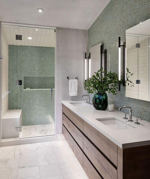 Contemporary double-vanity bathroom with a spacious glass-enclosed walk-in shower. The room features pale marble-look floor and shower-base surfaces, a green patterned tile feature wall extending behind the vanity and shower, a long white countertop with two integrated rectangular basins, and a wall-to-wall timber-look floating vanity with dark recessed pulls. Vertical illuminated mirrors and black fixtures provide contrast, while a green glass vase with leafy branches adds a natural accent.